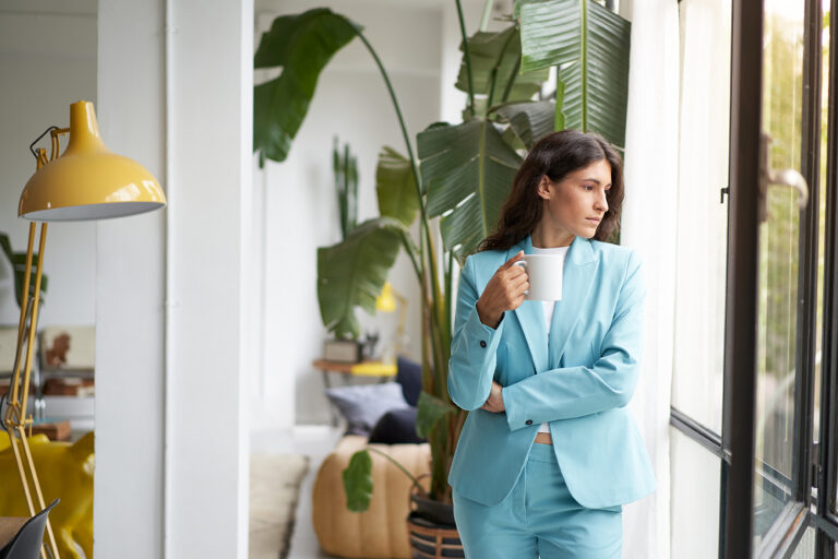 Woman in a light blue suit standing by a window holding a coffee mug, deep in thought, illustrating how it feels to suspect retaliation at work