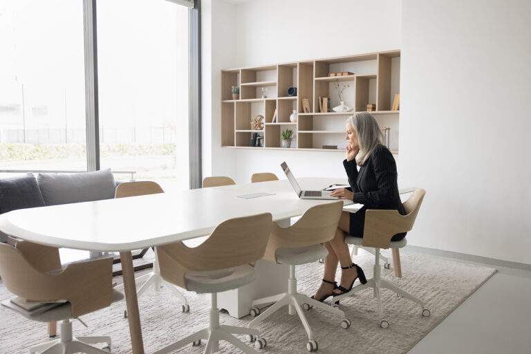 An older businesswoman with gray hair works alone at a laptop in an empty conference room, reflecting the isolation many employees over 40 feel when facing age discrimination at work.